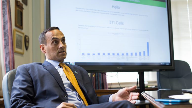 man in business suite sitting at table speaking, a large screen showing data is behind him